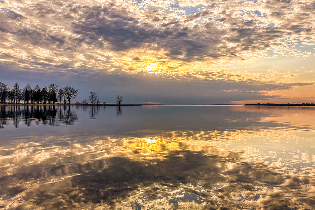 A serene sunset over the Ottawa River, where trees stand silhouetted against the sky’s vibrant hues, their reflections mirrored perfectly in the still water—nature's quiet masterpiece. Britannia Beach, Ottawa, Ontario