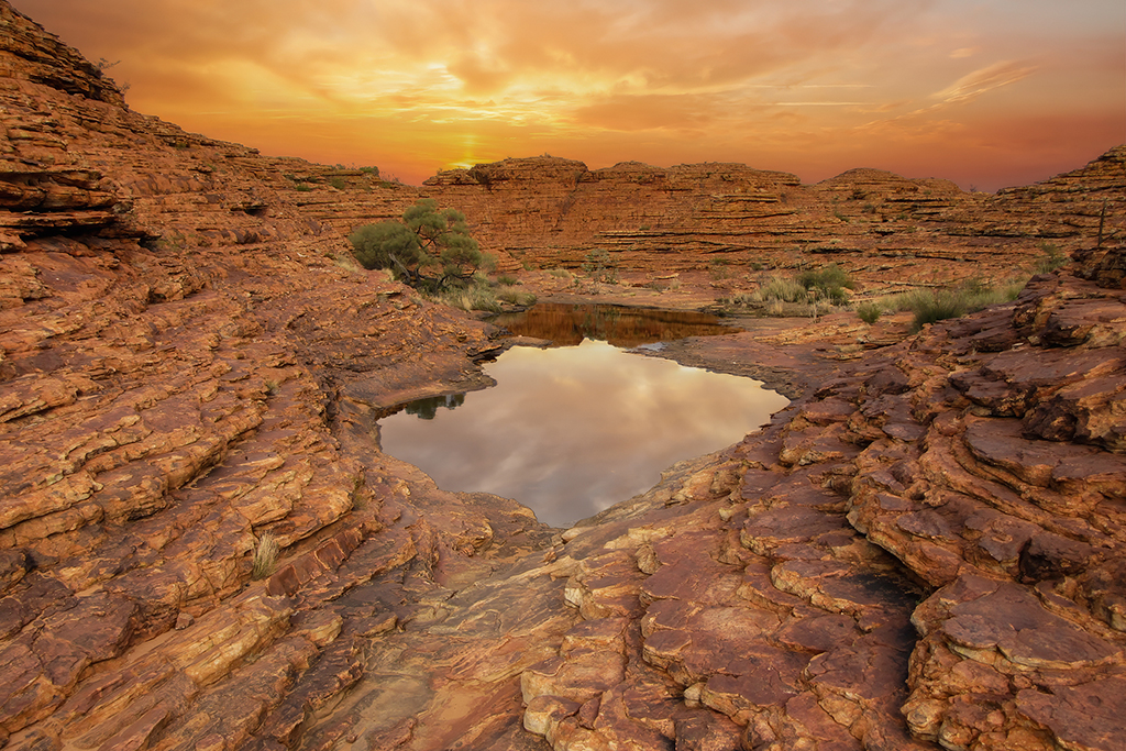 The golden hour ignites the red rock walls, as shadows deepen and a mirror-like pool reflects the drama overhead. King's Canyon, Australia